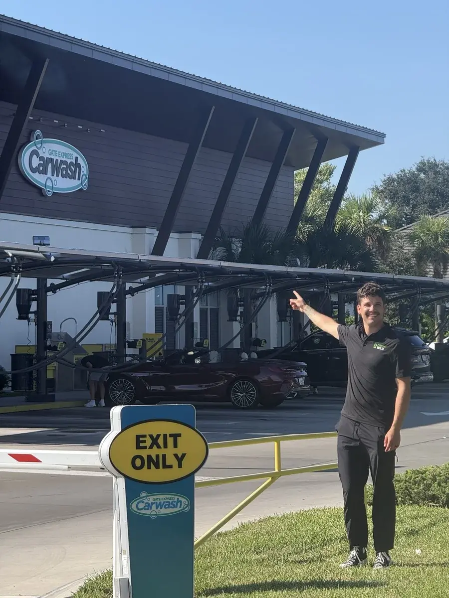 Kaden Ewald in a Grow Wild polo, pointing toward the GATE Express Carwash sign.